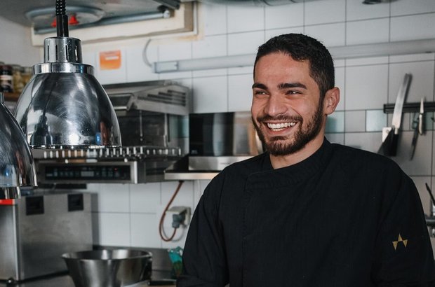 davit-kuechenchef Dunkelhaariger Mann in schwarzem Kochgewand lächelt in die Kamera, während er Gemüse in der Hotel-Küche schneidet.