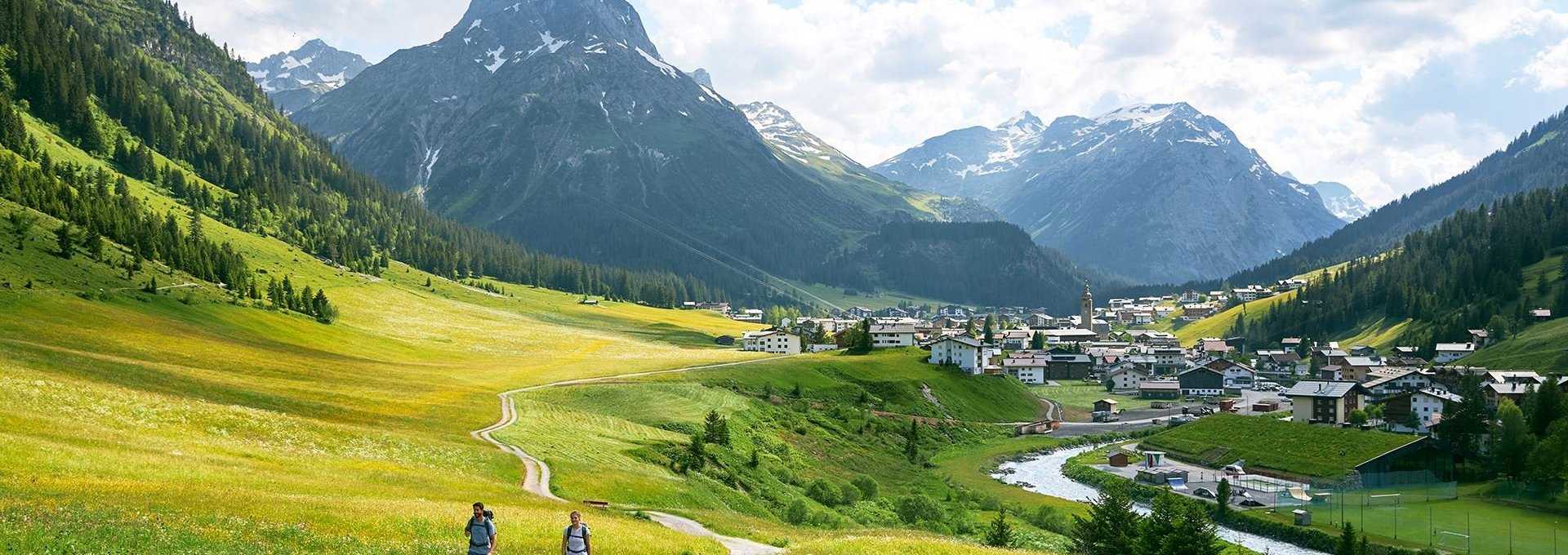 Lechweg mit Blick auf Lech Zwei Wanderer auf dem Lechweg. Wiesen sind neben dem Weg und im Hintergrund befinden sich die Berge.